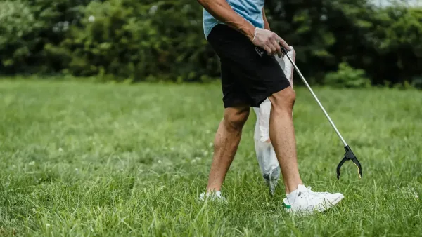 Man in shorts picking up litter off the grass with a long litter grabber.